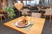 Ein elegant gedeckter Holztisch in einem stilvollen Restaurant mit einem angerichteten Teller, auf dem rosa gebratenes Fleisch, bunte Salate und Gemüse angerichtet sind. Im Hintergrund gemütliches Ambiente mit dekorativem Holzelement in Herzform und Bergb
