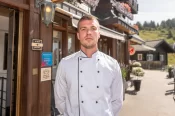 Ein junger Koch in weißer Kochjacke steht vor dem Eingang eines Hotels oder Restaurants. Im Hintergrund sind Auszeichnungen an der Wand angebracht sowie ein traditionell gestaltetes Gebäude mit Terrasse und Blumenschmuck in alpiner Umgebung.