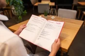 Person sitzt an einem Holztisch in einem stilvoll eingerichteten Restaurant und hält eine geöffnete Speisekarte des Restaurants Bettmerhof | Pizza Bello in den Händen. Auf dem Tisch stehen zwei leere Weingläser und eine kleine Zimmerpflanze.